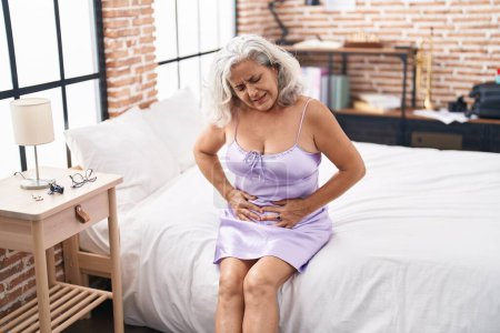 Téléchargez les photos : Femme aux cheveux gris d'âge moyen souffrant de douleurs menstruelles assise sur le lit dans la chambre - en image libre de droit