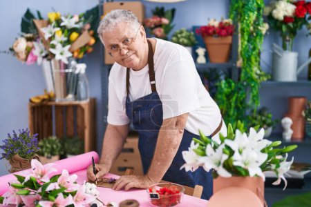 Foto de Florista hombre de pelo gris de mediana edad sonriendo escritura segura en carta sobre en floristería - Imagen libre de derechos