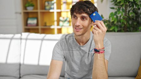 Téléchargez les photos : Jeune homme hispanique tenant une carte de crédit assis sur le canapé pensant à la maison - en image libre de droit