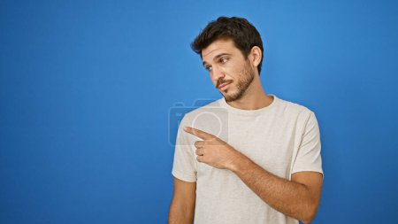 Photo for Handsome young hispanic man with beard pointing to his left against a solid blue outdoor background. - Royalty Free Image