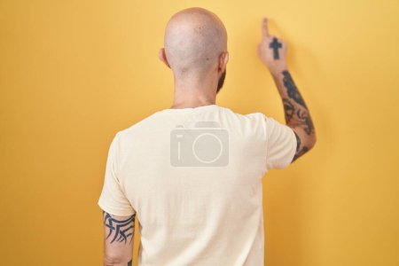 Foto de Hombre hispano con tatuajes de pie sobre fondo amarillo posando hacia atrás apuntando hacia adelante con la mano del dedo - Imagen libre de derechos