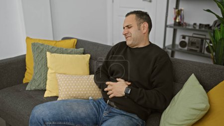 Junger hispanischer Mann mit Schmerzen, der seinen Bauch umklammert, während er drinnen auf einer grauen Couch mit bunten Kissen sitzt.