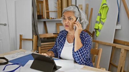 Photo for Mature woman on phone multitasking in workshop with tablet and blueprints. - Royalty Free Image