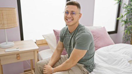 Photo for Smiling young hispanic man sitting on a bed in a well-lit bedroom interior, exuding casual charm and confidence. - Royalty Free Image