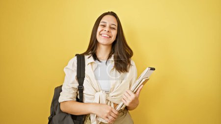 Photo for Smiling, young and beautiful hispanic woman student with cool confidence, book in hand and backpack, posing on an isolated yellow background, enjoying a fun and positive lifestyle - Royalty Free Image