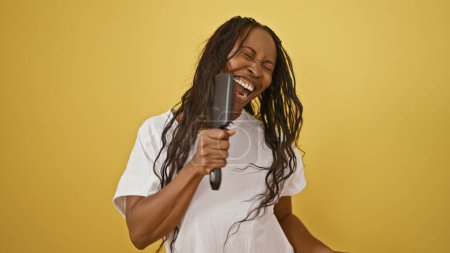 Téléchargez les photos : Une femme afro-américaine joyeuse chantant avec une brosse à cheveux sur un fond jaune. - en image libre de droit