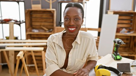 Foto de Sonriente mujer afroamericana carpintero sentado con confianza en la mesa de carpintería en su taller ocupado, interior - Imagen libre de derechos