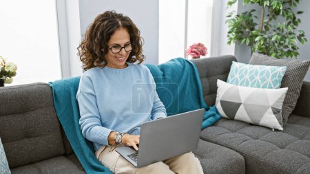 Foto de Mujer hispana usando el portátil cómodamente en su moderna sala de estar en casa - Imagen libre de derechos