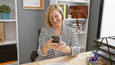 Photo for A cheerful middle-aged blond woman enjoys using her smartphone in a modern office setting - Royalty Free Image
