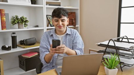 Photo for Young man smiles while using smartphone at office desk with laptop and documents. - Royalty Free Image
