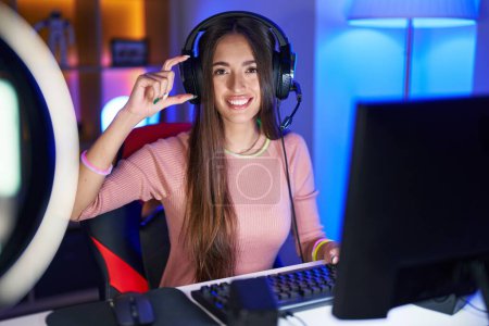 Photo for Young hispanic woman playing video games smiling and confident gesturing with hand doing small size sign with fingers looking and the camera. measure concept. - Royalty Free Image