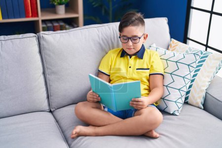 Photo for Adorable hispanic boy reading book sitting on sofa at home - Royalty Free Image