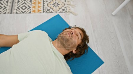 Photo for A relaxed young caucasian man lies with closed eyes on a blue yoga mat in a bright home interior. - Royalty Free Image