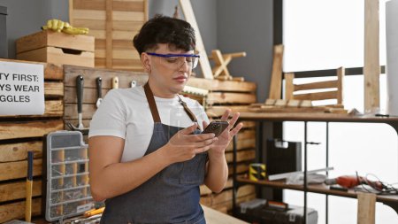 Photo for A young man wearing safety glasses reviews a message on his phone in a well-organized carpentry workshop - Royalty Free Image