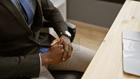 Foto de Hombre afroamericano en un traje de negocios sentado en un escritorio de oficina con una computadora portátil en el fondo. - Imagen libre de derechos