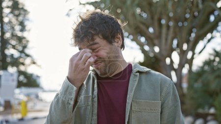 Distressed hispanic man pinching the bridge of his nose outdoors against a park backdrop.