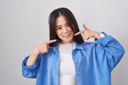 Téléchargez les photos : Jeune femme chinoise debout sur fond blanc souriant gai montrant et pointant avec les doigts dents et la bouche. concept de santé dentaire. - en image libre de droit