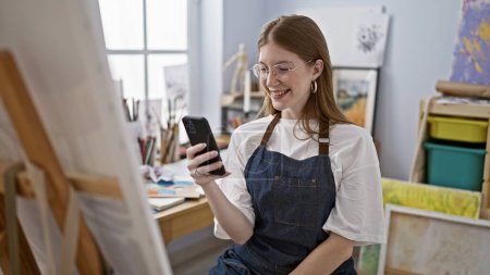 Téléchargez les photos : Jeune artiste blonde utilisant smartphone souriant au studio d'art - en image libre de droit