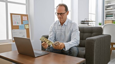 Foto de Hombre de mediana edad seguro, un trabajador de negocios feliz, disfrutando de su éxito mientras se sienta en el interior de la oficina, contando dólares con una sonrisa en su computadora portátil - Imagen libre de derechos