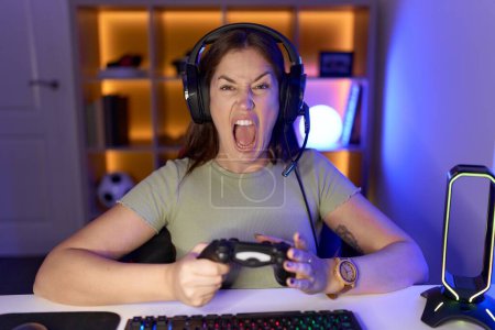 Téléchargez les photos : Belle femme brune jouant à des jeux vidéo portant des écouteurs en colère et fous hurlant frustrés et furieux, criant de colère. rage et concept agressif. - en image libre de droit