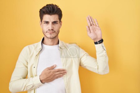 Photo for Young hispanic man standing over yellow background swearing with hand on chest and open palm, making a loyalty promise oath - Royalty Free Image