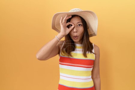 Téléchargez les photos : Femme chinoise du Moyen Âge portant un chapeau d'été sur fond jaune faisant un geste correct choqué par le visage surpris, les yeux regardant à travers les doigts. expression incrédule. - en image libre de droit