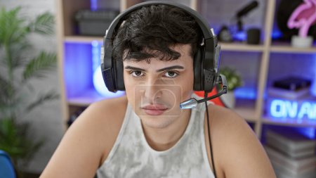 Foto de Hombre joven con auriculares en una sala de juegos mirando a la cámara, iluminado por luces de neón en casa. - Imagen libre de derechos