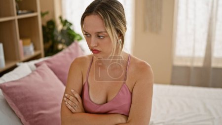 Foto de Mujer rubia joven sentada en la cama con expresión infeliz y brazos cruzados gesto en el dormitorio - Imagen libre de derechos