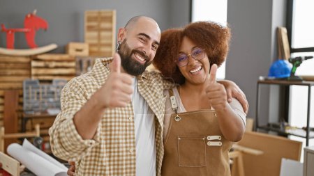 Photo for Two carpentry partners beaming, giving okay sign while hugging inside their woodworking workshop - Royalty Free Image