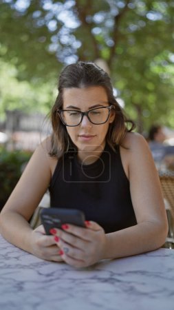 Photo for Smiling beautiful hispanic woman, cheerfully using smartphone while sitting at cafe's outdoor terrace, enjoying a coffee, connection with the digital world enhancing her happiness. - Royalty Free Image