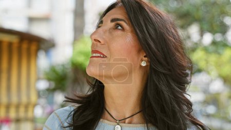 A thoughtful hispanic woman in her mature years gazes up in a lush garden setting, epitomizing serenity and outdoor grace.