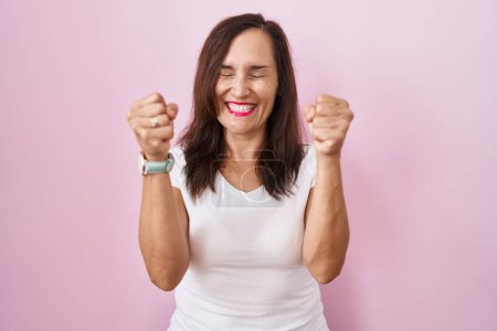Téléchargez les photos : Femme brune du moyen âge debout sur fond rose excité pour le succès avec les bras levés et les yeux fermés célébrant la victoire en souriant. concept gagnant. - en image libre de droit