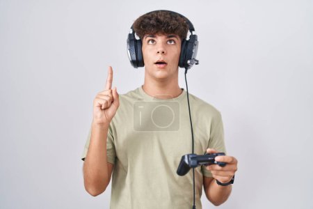 Téléchargez les photos : Adolescent hispanique jouant contrôleur de tenue de jeu vidéo étonné et surpris regardant vers le haut et pointant avec les doigts et les bras levés. - en image libre de droit