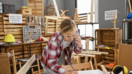 Foto de Una joven carpintera enfocada en atuendos casuales está en una llamada telefónica mientras escribe notas en un taller de carpintería. - Imagen libre de derechos