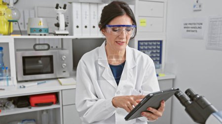 Foto de Científica madura en bata de laboratorio usando tableta en ambiente de laboratorio, retratando la investigación de la salud. - Imagen libre de derechos