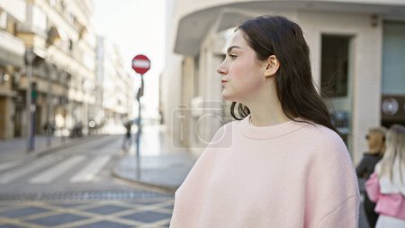 Foto de Mujer caucásica joven en traje casual de pie pensativamente en una calle urbana soleada con edificios y una señal de stop en el fondo. - Imagen libre de derechos