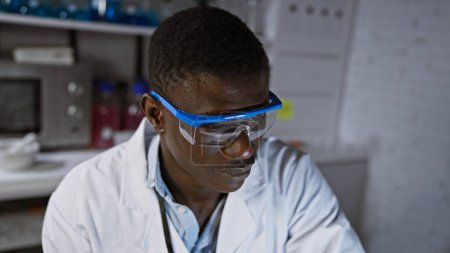 Photo for An african man wearing safety glasses in a laboratory setting, deep in thought or analysis. - Royalty Free Image