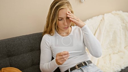 Young caucasian woman looking concerned at thermometer while sitting on a couch indoors.