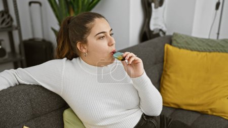 Téléchargez les photos : Jeune femme hispanique se détendre sur le canapé dans le salon moderne tout en vaping - en image libre de droit