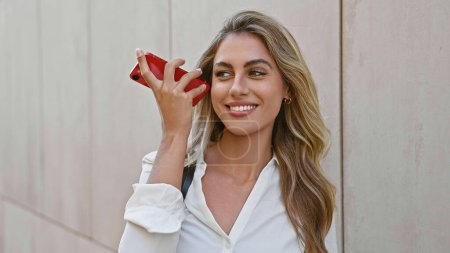 Téléchargez les photos : Une jeune femme blonde confiante écoutant joyeusement un message vocal sur son smartphone, debout dans une rue animée de la ville, souriant d'une oreille à l'autre. - en image libre de droit