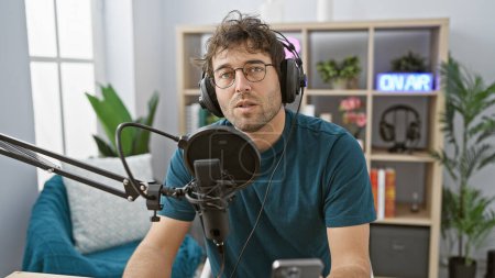 Ein junger hispanischer Mann mit Bart spricht in einem Radiostudio-Set mit Kopfhörern in ein Mikrofon und deutet auf eine Übertragungsszene im Innenraum hin..