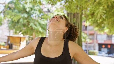 Téléchargez les photos : Femme hispanique aux cheveux bouclés jouit de la nature dans un parc urbain ensoleillé, émanant la jeunesse et la beauté. - en image libre de droit