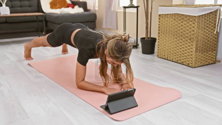 Photo for Sporty, attractive blonde woman getting her abs burn with focused indoor core exercise training - streaming online fitness workout on laptop from comfort of her living room - Royalty Free Image