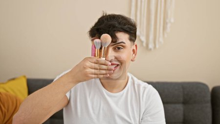 Photo for A handsome young man smiles while holding makeup brushes in a modern living room, representing beauty and lifestyle themes. - Royalty Free Image