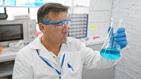 Téléchargez les photos : Un homme mûr scientifique examine un liquide bleu dans une fiole dans un cadre de laboratoire portant des lunettes de protection. - en image libre de droit