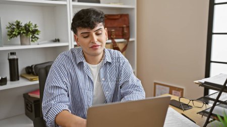 Téléchargez les photos : Un jeune homme travaille attentivement sur un ordinateur portable dans un cadre de bureau moderne, avec des étagères et un décor en arrière-plan. - en image libre de droit