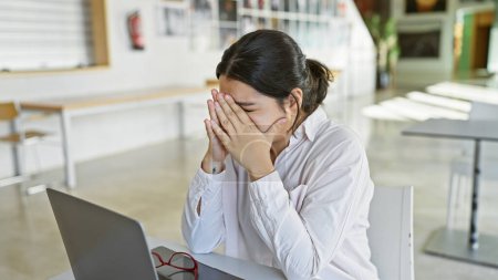Photo for A stressed hispanic woman in a white shirt covers her face with her hands at a modern office table, with a laptop and glasses nearby. - Royalty Free Image