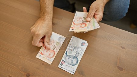 Téléchargez les photos : Homme comptant les dollars singapouriens à une table en bois à l'intérieur, dépeignant la finance et le cadre domestique. - en image libre de droit