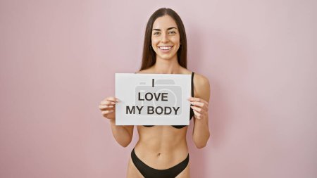 Photo for Beautiful young hispanic woman in lingerie, confidently enjoying her sensual morning, proudly holds 'i love my body' banner over isolated pink background, spreading a joyful, body-positive message. - Royalty Free Image