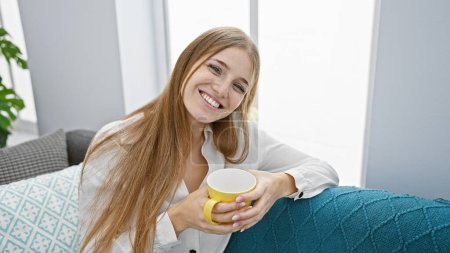 Téléchargez les photos : Une jeune femme souriante tenant une tasse, assise dans un salon moderne et lumineux. - en image libre de droit
